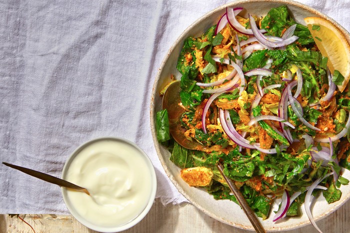 A bowl filled with green sprout tops and topped with sliced red onion, green coriander with yogurt on the side