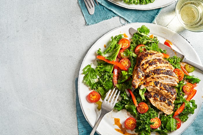 A grey background with two white plates topped with green salad leaves, halved tomatoes and slices of cooked chicken