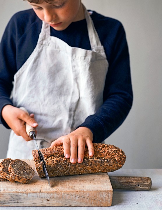 A person wearing an apron slicing a loaf of seeded bread