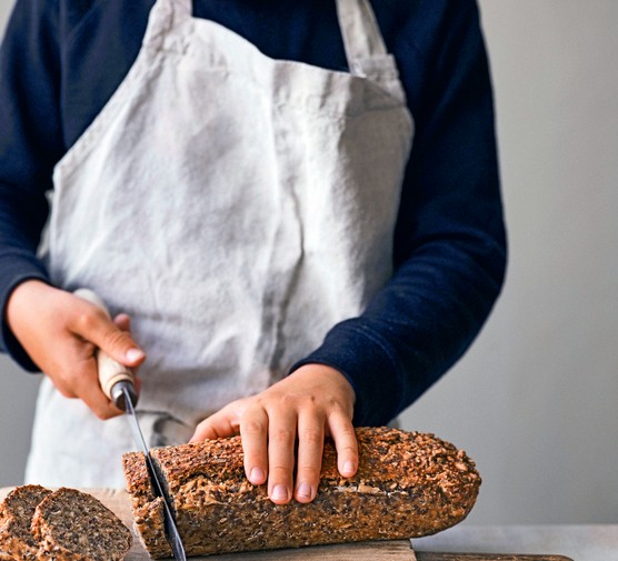 A person wearing an apron slicing a loaf of seeded bread