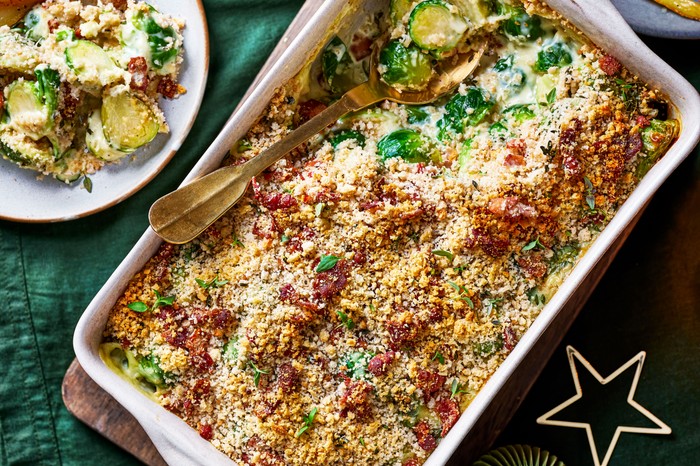 A brussels sprout and bacon gratin in a large white baking dish, next to a plate of parsnips
