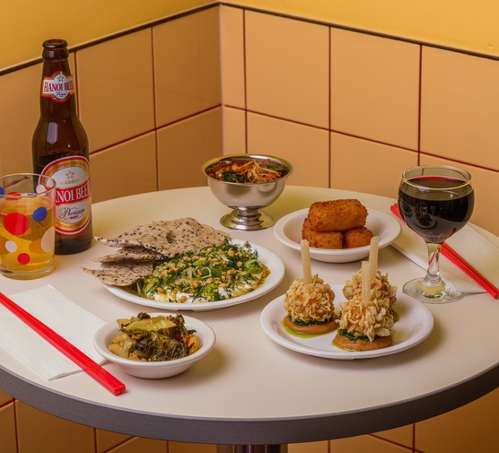 A selection of small plates, beer and wine on a bar table, with a butter yellow tiled wall in the background