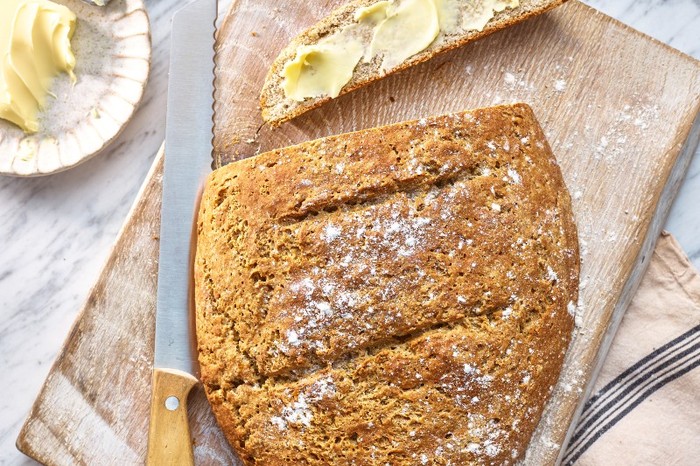 A loaf of bread on a wooden board with a slice cut out of the end and spread with butter