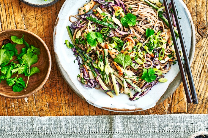 A wooden table topped with a noodle and shredded veg salad, with a pair of black chopsticks on the side