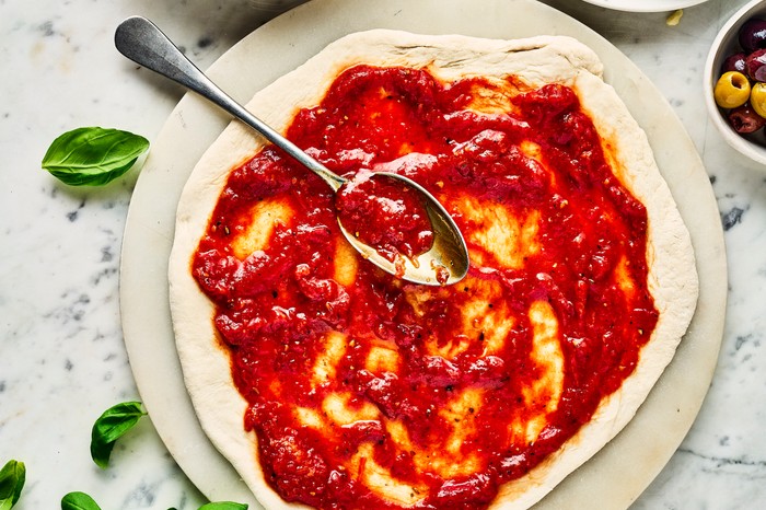 An uncooked pizza base with tomato sauce spread over it, next to a plate of grated cheese, bowl of more sauce and some fresh basil leaves