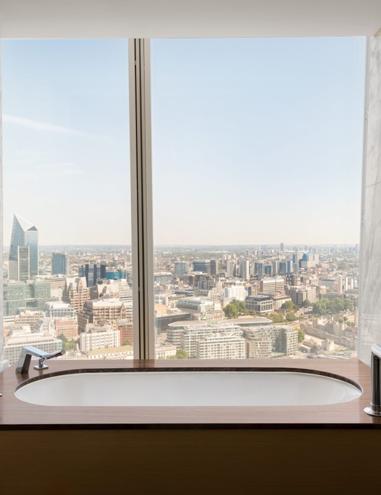 A bathtub in a hotel room which has a glass window looking out over London