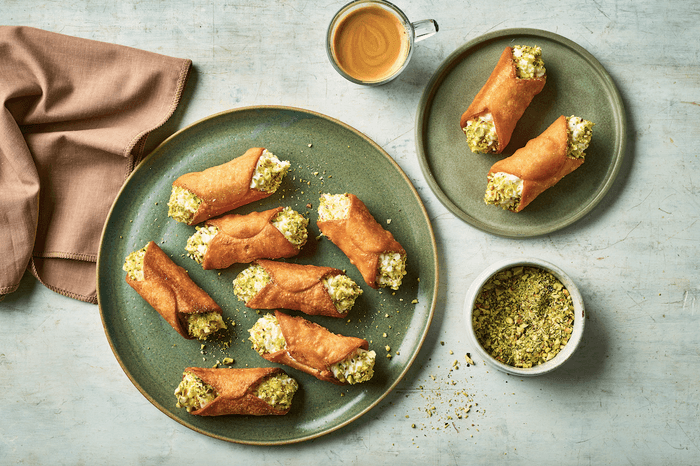 A large plate of cannoli on a grey background with a shot of espresso and a small bowl of chopped pistachios